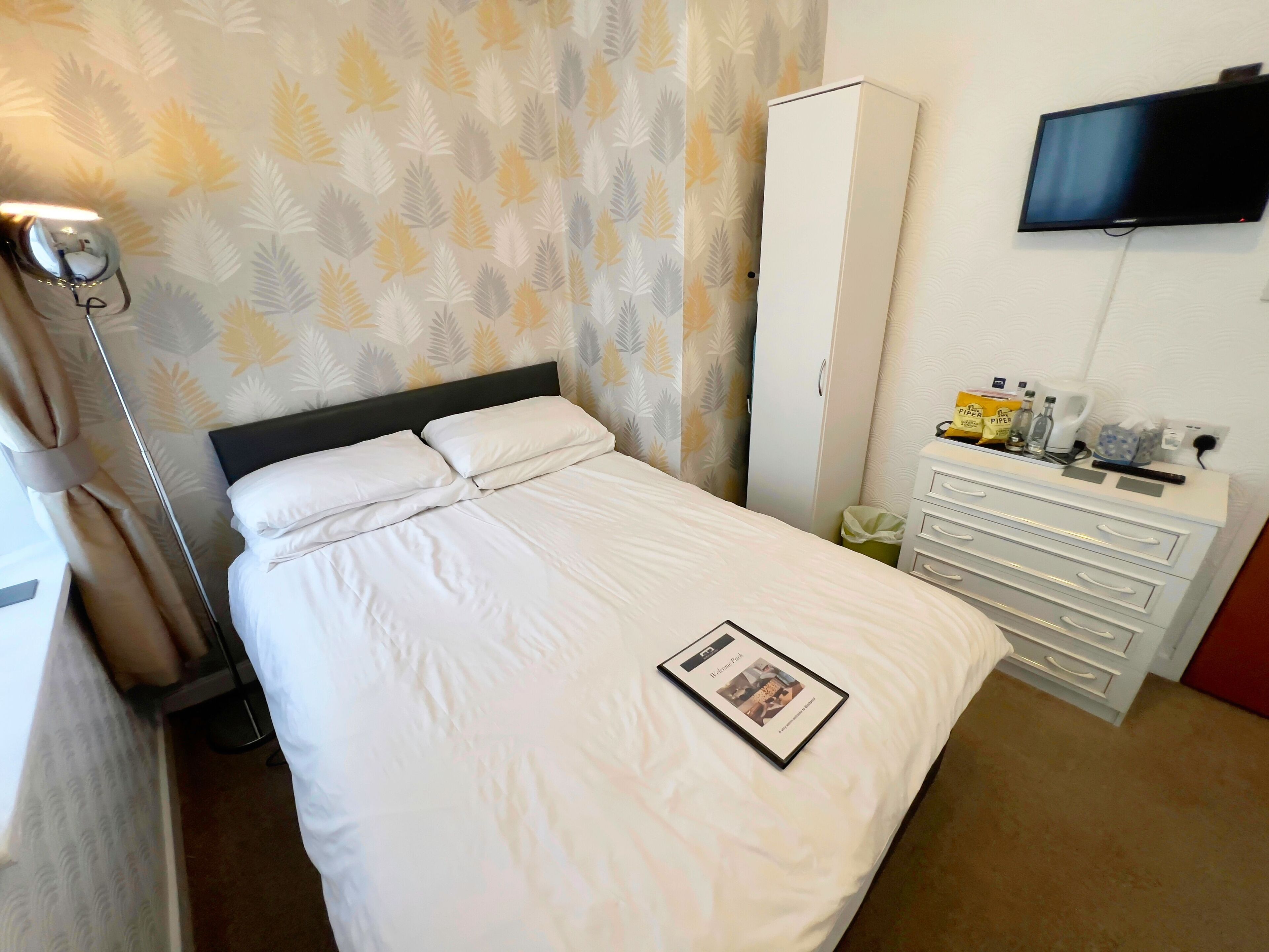 A neatly made bed with white linens, a bedside table with a remote control, a television mounted on the wall, and a wardrobe with a mirror.