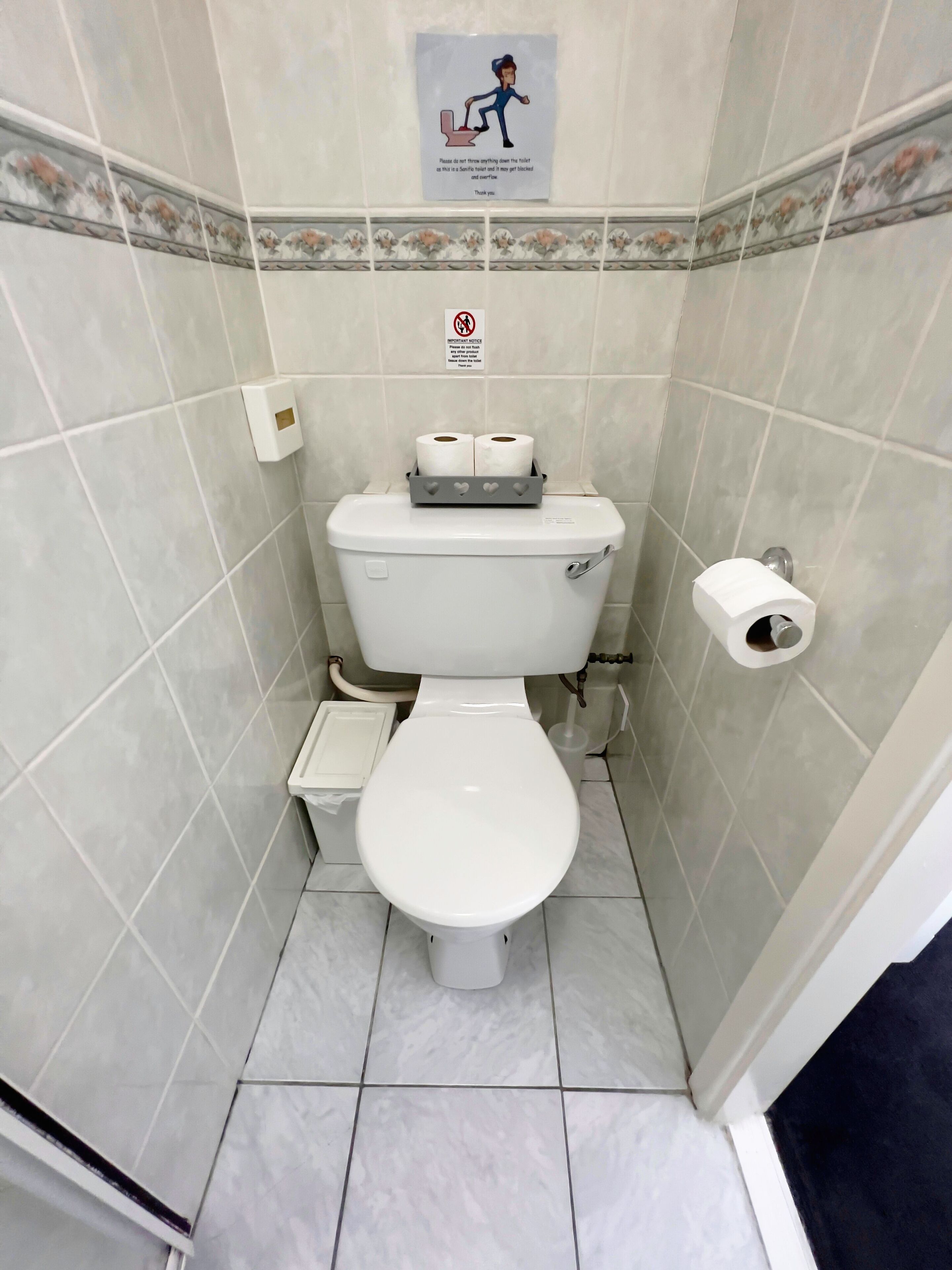 A small, tiled bathroom with a toilet, a paper towel holder, and a toilet paper roll.