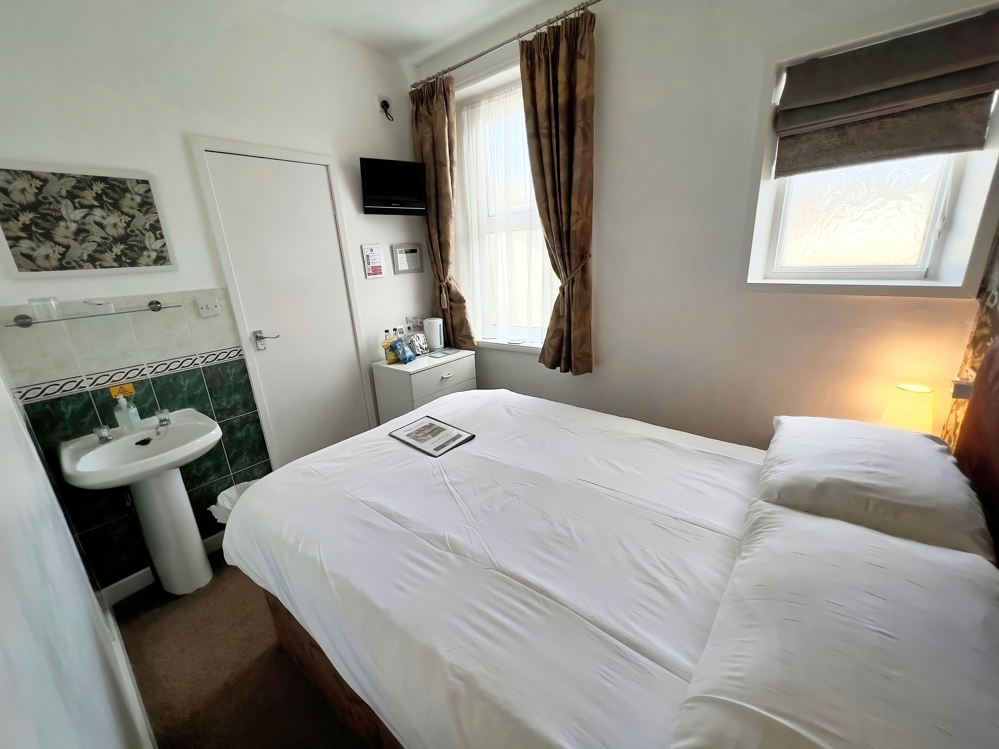 Een hotelkamer met een bed, nachtkastje, televisie, raam met gordijnen, wastafel en spiegel.