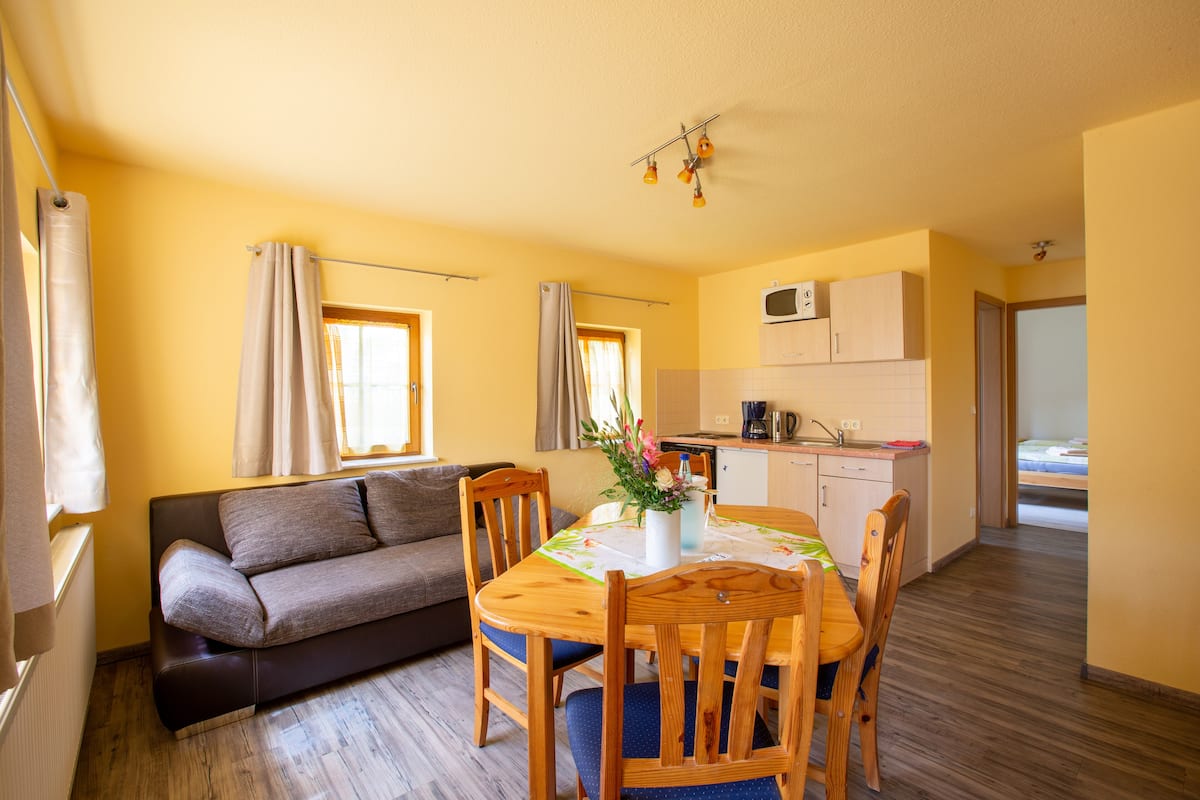 Un espace de vie compact comprenant un canapé, une table à manger, des chaises, un coin cuisine et une petite chambre.