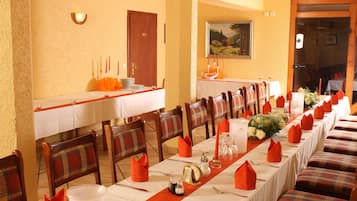 A dining area with long tables set for multiple guests, wooden chairs with plaid upholstery, and a warm-toned wall.