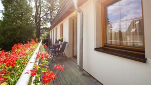 Un patio techado al aire libre con una mesa y sillas, una ventana con vista a un edificio y una baranda adornada con flores rojas.