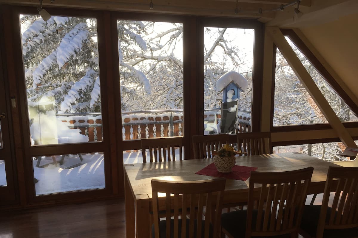 Une table de salle à manger en bois, entourée de chaises, un vase de fleurs et une vue sur des arbres couverts de neige, visible à travers de grandes fenêtres.