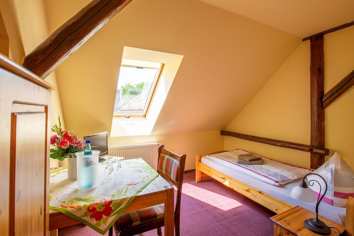Une petite pièce agréable, dotée d’un plafond en pente, d’un lit simple, d’une table en bois avec une chaise, d’une fenêtre offrant une vue et d’un radiateur.