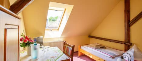 Una habitación pequeña y acogedora con techo inclinado, una cama individual, una mesa de madera con una silla, un ventanal con vistas y un radiador.