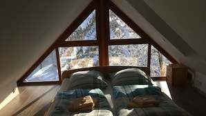 Un dormitorio con techo inclinado, dos camas con ropa de cama escocesa y vistas a árboles nevados a través de amplias ventanas triangulares.