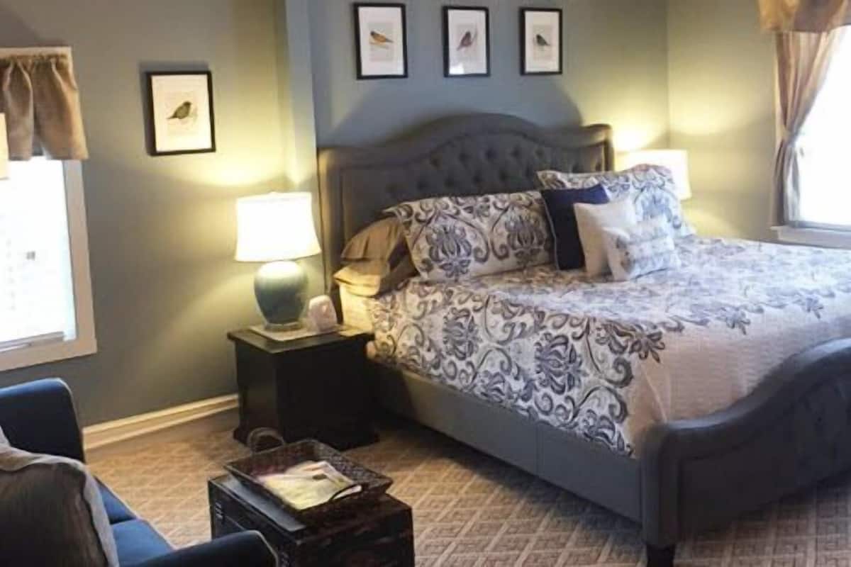 A bedroom with a bed, two bedside tables, a lamp, a chair, and a window with curtains.
