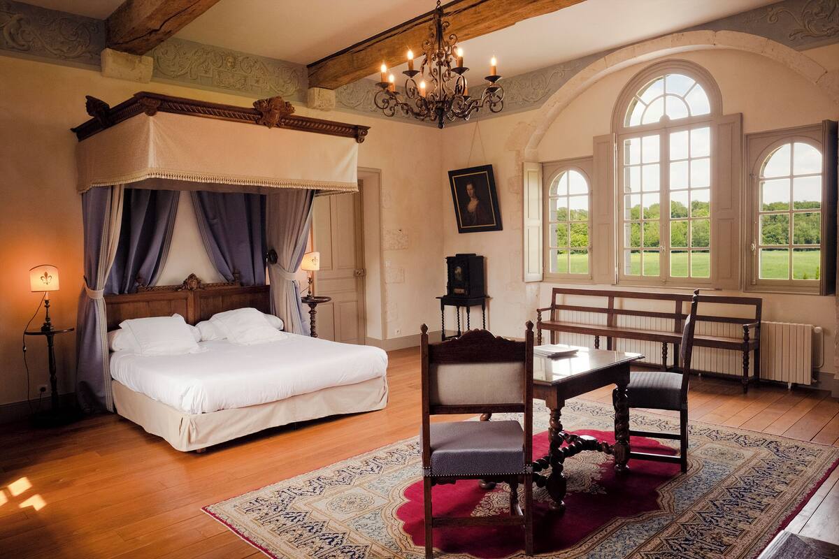 Quarto com cama de dossel, lustre, mesa de jantar com cadeiras e grandes janelas com vista para o verde.