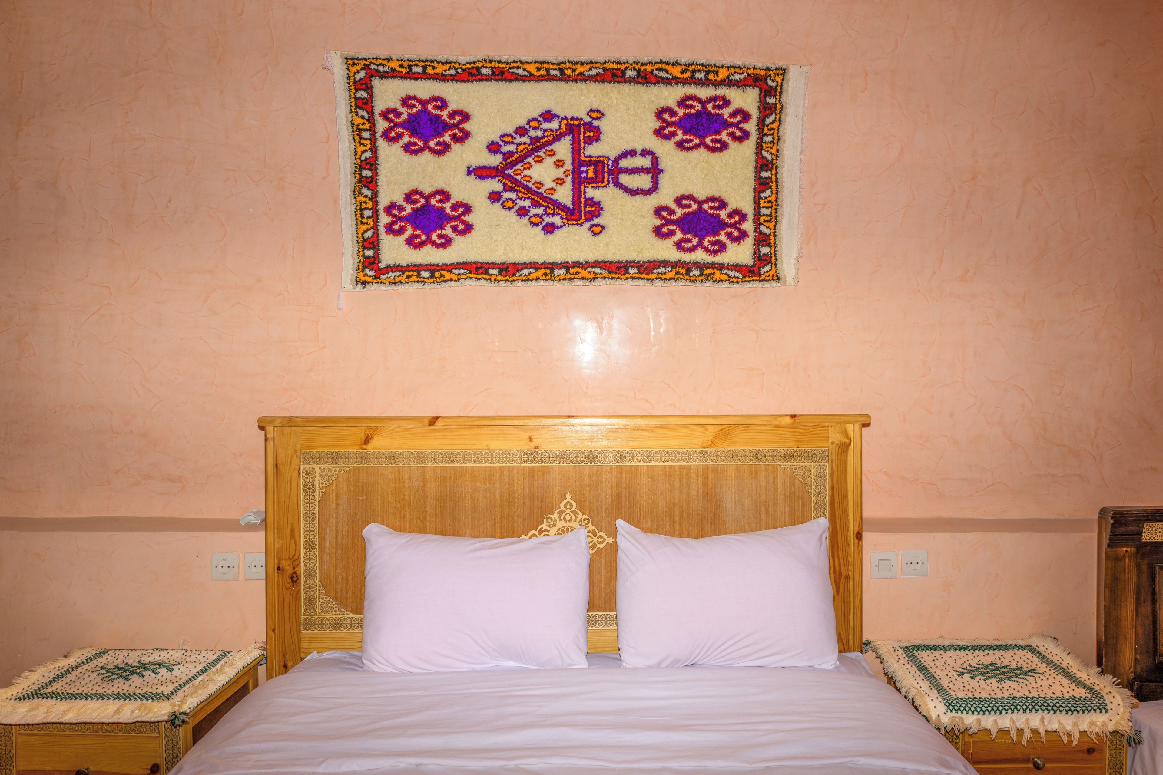 A bedroom with a wooden bed, two pillows, a decorative wall hanging, and two bedside tables with patterned tablecloths.