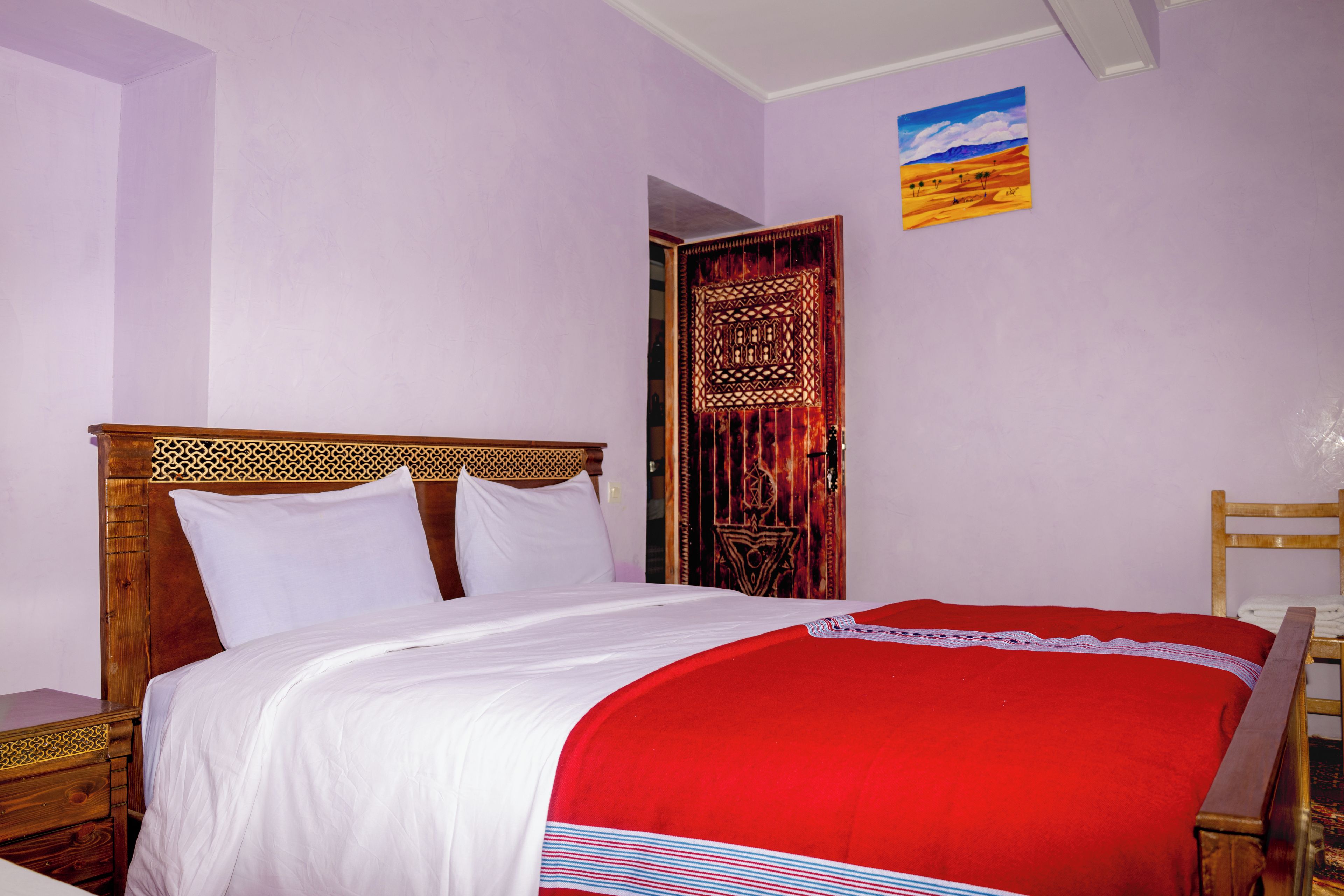 A room with a bed, a wooden headboard, a nightstand, a red and white striped blanket, and a door with intricate patterns.