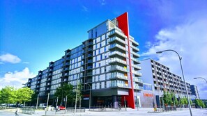 A modern multi-story building with a prominent red vertical element, large windows, and a visible storefront with signage. - City Apartments Milton Keynes Vizion (Milton Keynes)