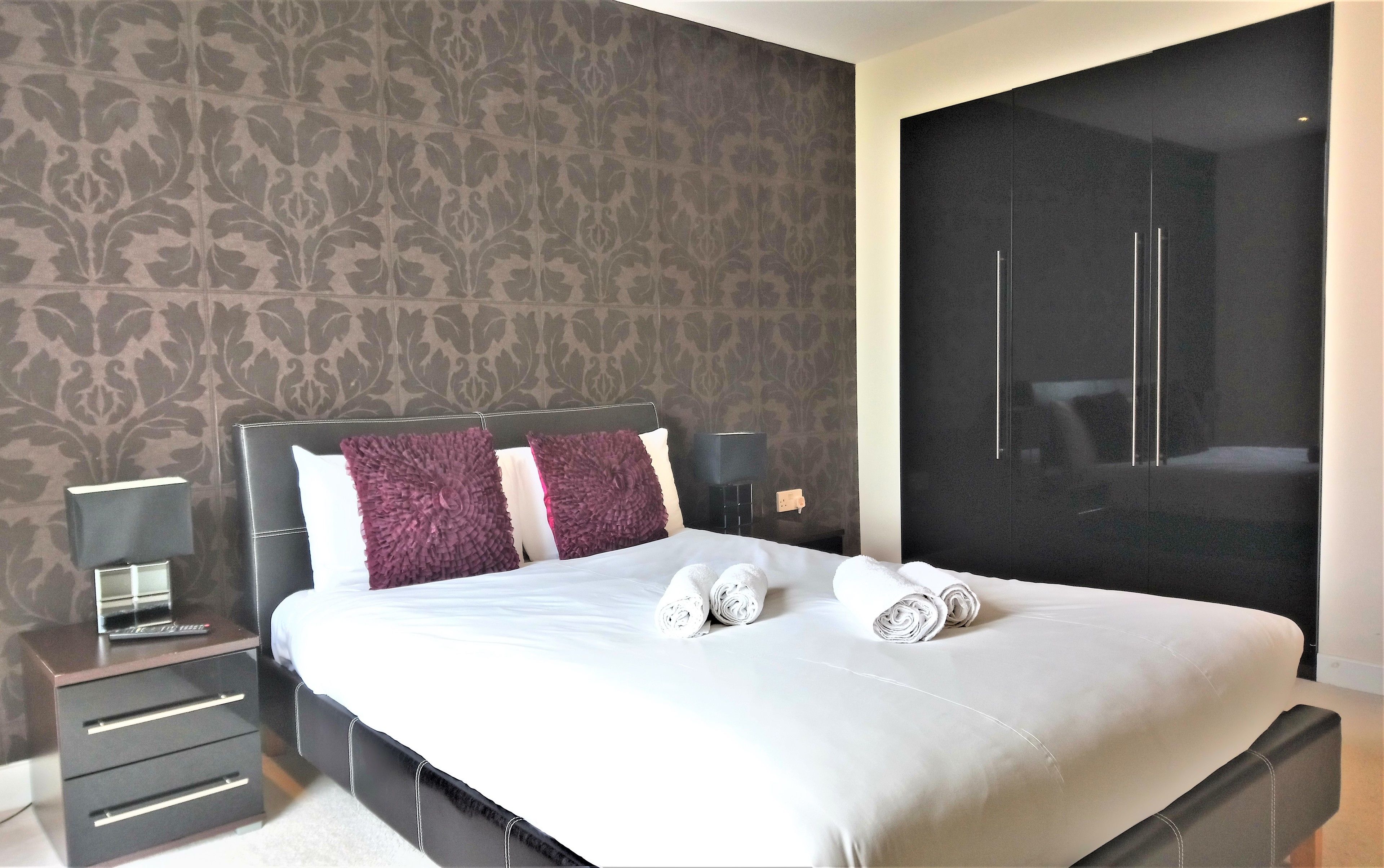 A neatly made bed with white and purple pillows, a bedside table with a lamp, and a large wardrobe.