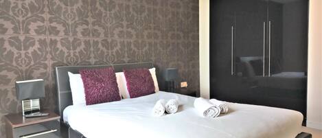 A neatly made bed with white and purple pillows, a bedside table with a lamp, and a large wardrobe.
