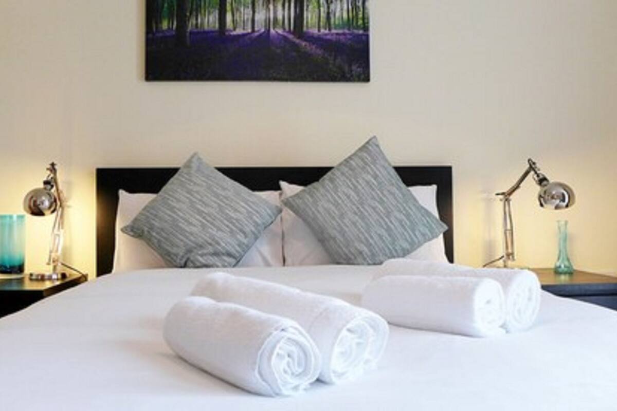 A neatly made bed with two pillows, a pair of rolled towels, and a painting above it.