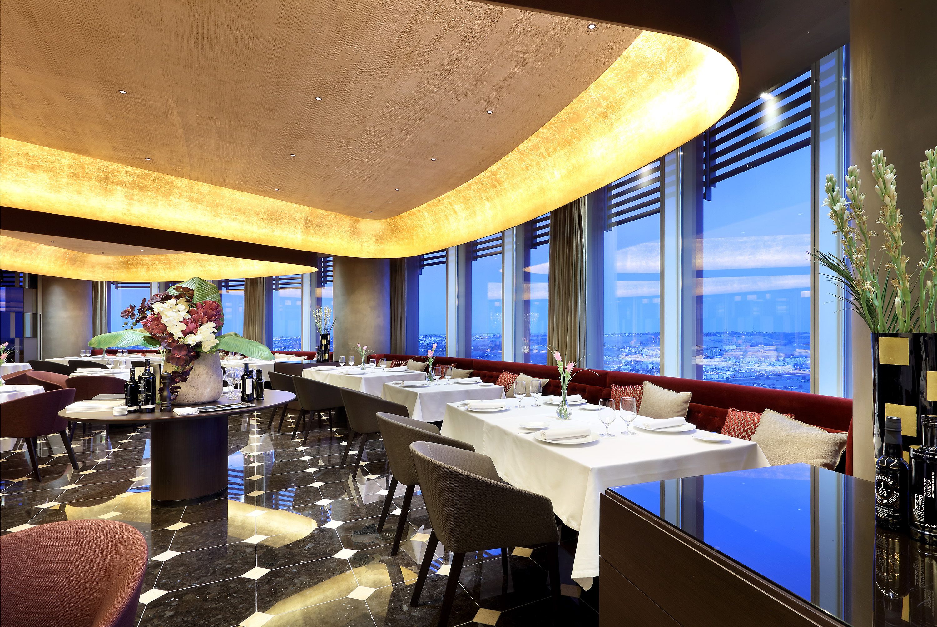 A restaurant with tables set for dining, a large window offering a city view, and a modern ceiling design.