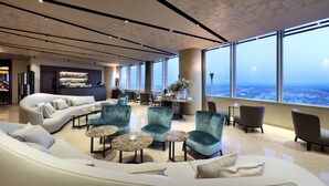 A modern lounge area with a curved sofa, armchairs, and round tables. A bar with a display of bottles is visible in the background. - Eurostars Torre Sevilla (Seville)