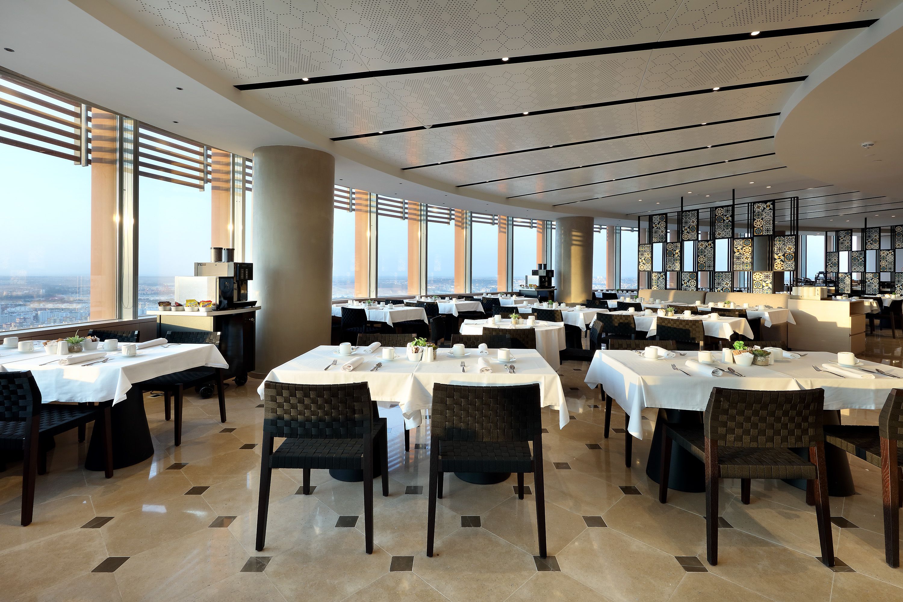 A spacious dining area with multiple tables set for guests, large windows offering a city view, and a modern ceiling design.