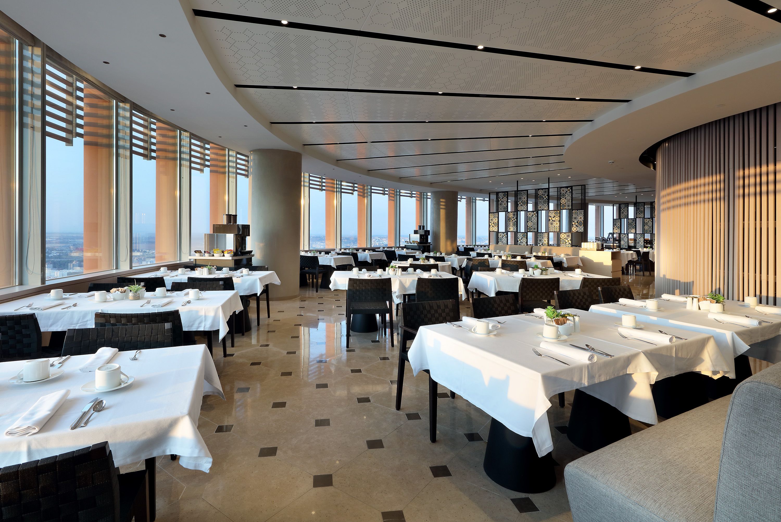 Un vaste espace dédié à la salle à manger, avec de nombreuses tables dressées, de grandes fenêtres offrant une vue sur la ville et un design moderne au niveau du plafond.