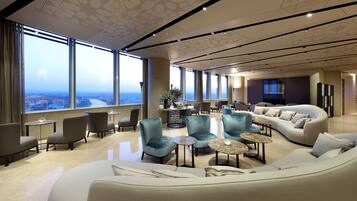 A spacious lounge area with a large window offering a city view, comfortable seating, and a modern ceiling design.