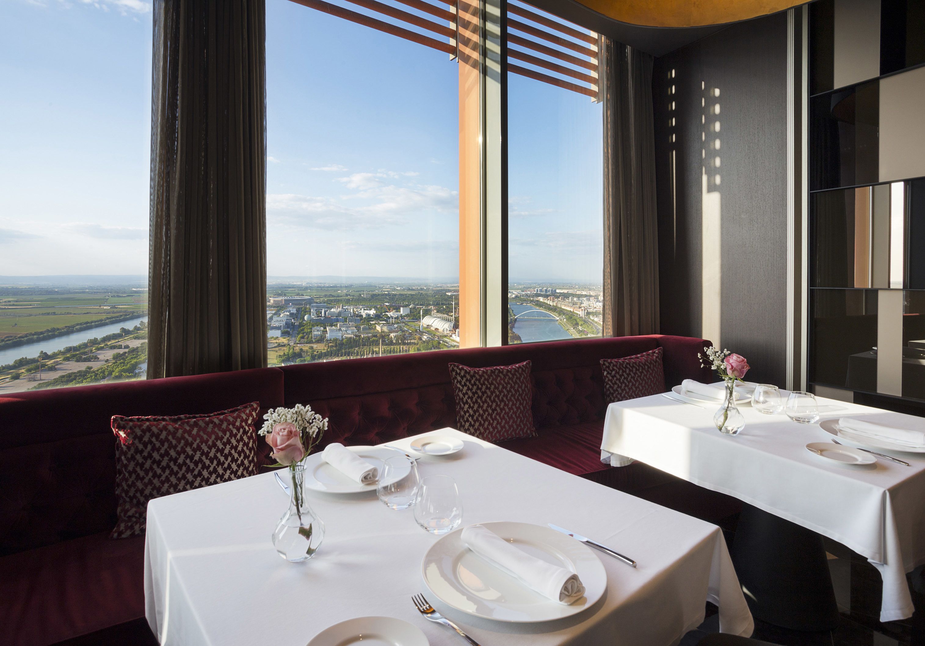 Un restaurante con mesas listas para comer, amplios ventanales que ofrecen vistas a un paisaje urbano, y un banco tapizado en rojo.