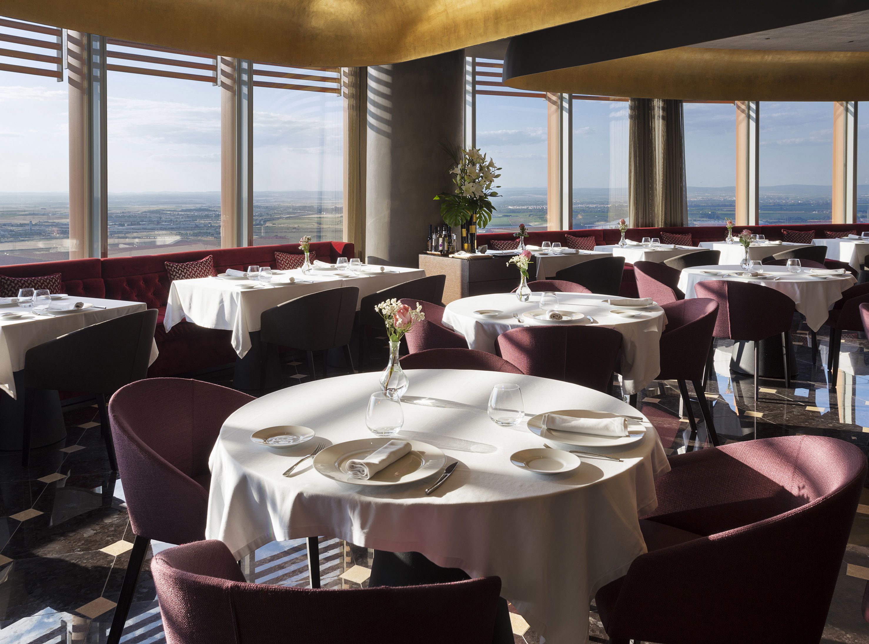 Een restaurant met ronde tafels, rode bekleden zitjes en grote ramen met uitzicht over de stad.
