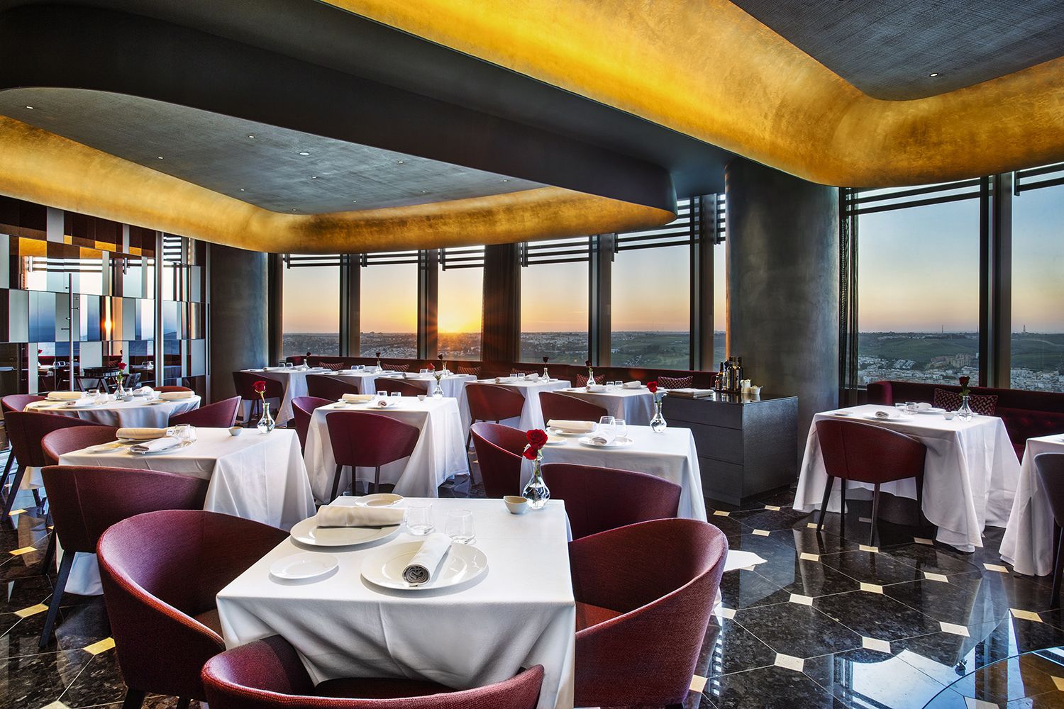 Un restaurante con mesas preparadas para comer, grandes ventanales que ofrecen vistas a la ciudad y una decoración interior moderna.