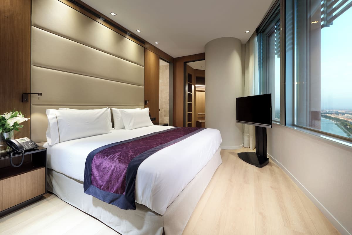 Habitación de hotel moderna con una cama grande, una mesita de noche con teléfono, televisión y vistas a la ciudad a través de amplias ventanas.