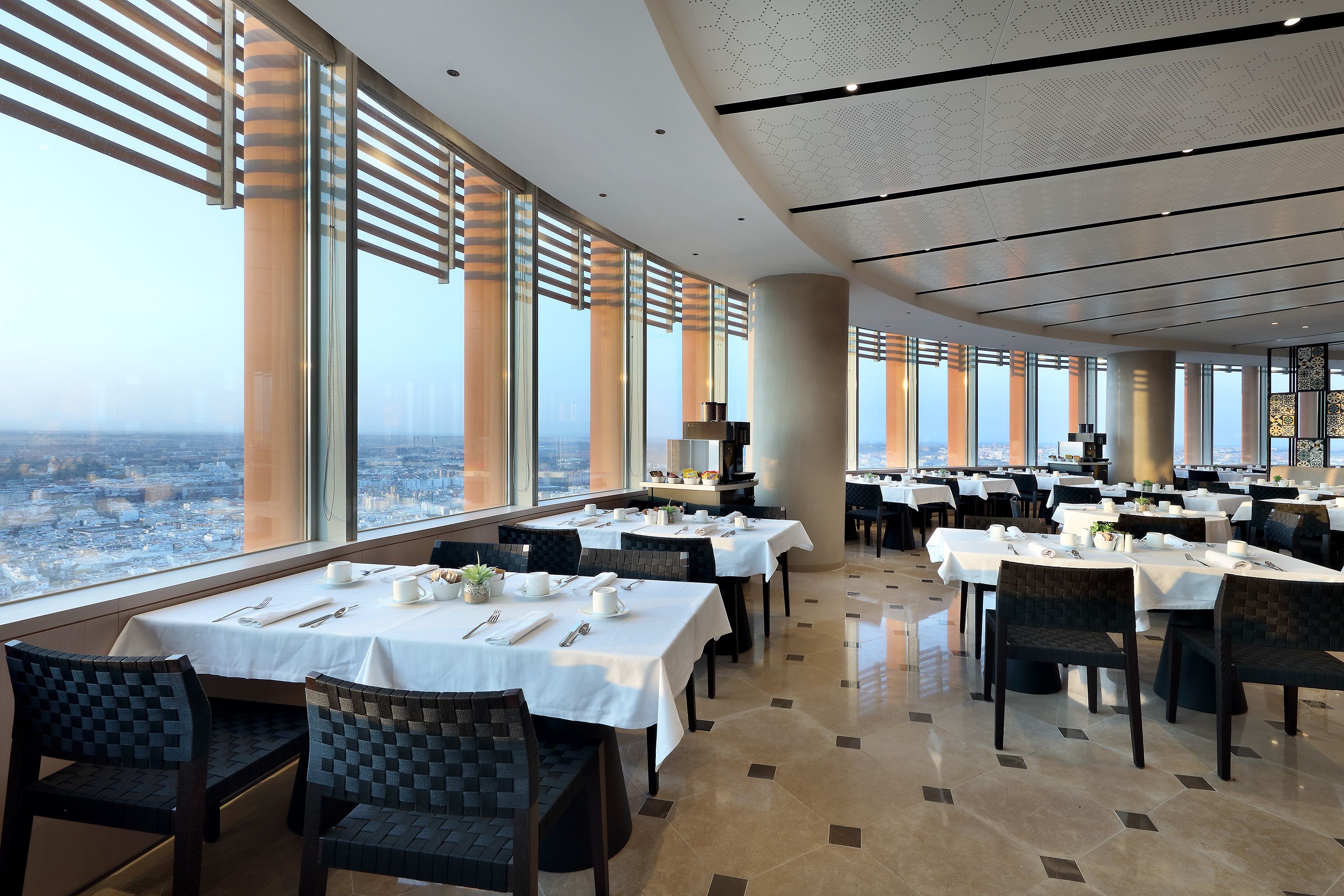 Un vaste espace dédié à la salle à manger, avec des tables dressées pour le service, de grandes fenêtres offrant une vue sur la ville et un design moderne au niveau du plafond.