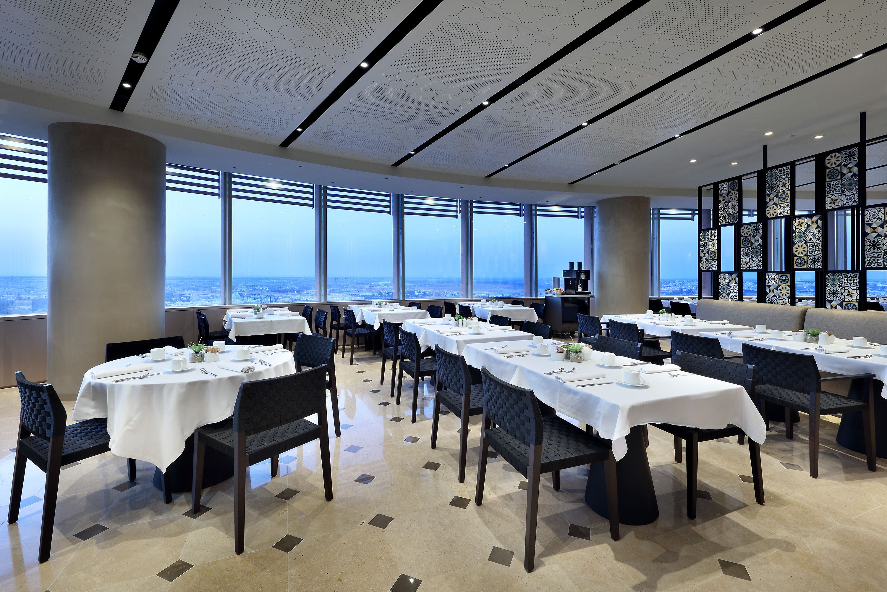 Un comedor con mesas dispuestas para el servicio, ventanas amplias con vistas a la ciudad y una pared decorativa con paneles estampados.
