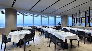 Un comedor con mesas dispuestas para el servicio, ventanas amplias con vistas a la ciudad y una pared decorativa con paneles estampados.