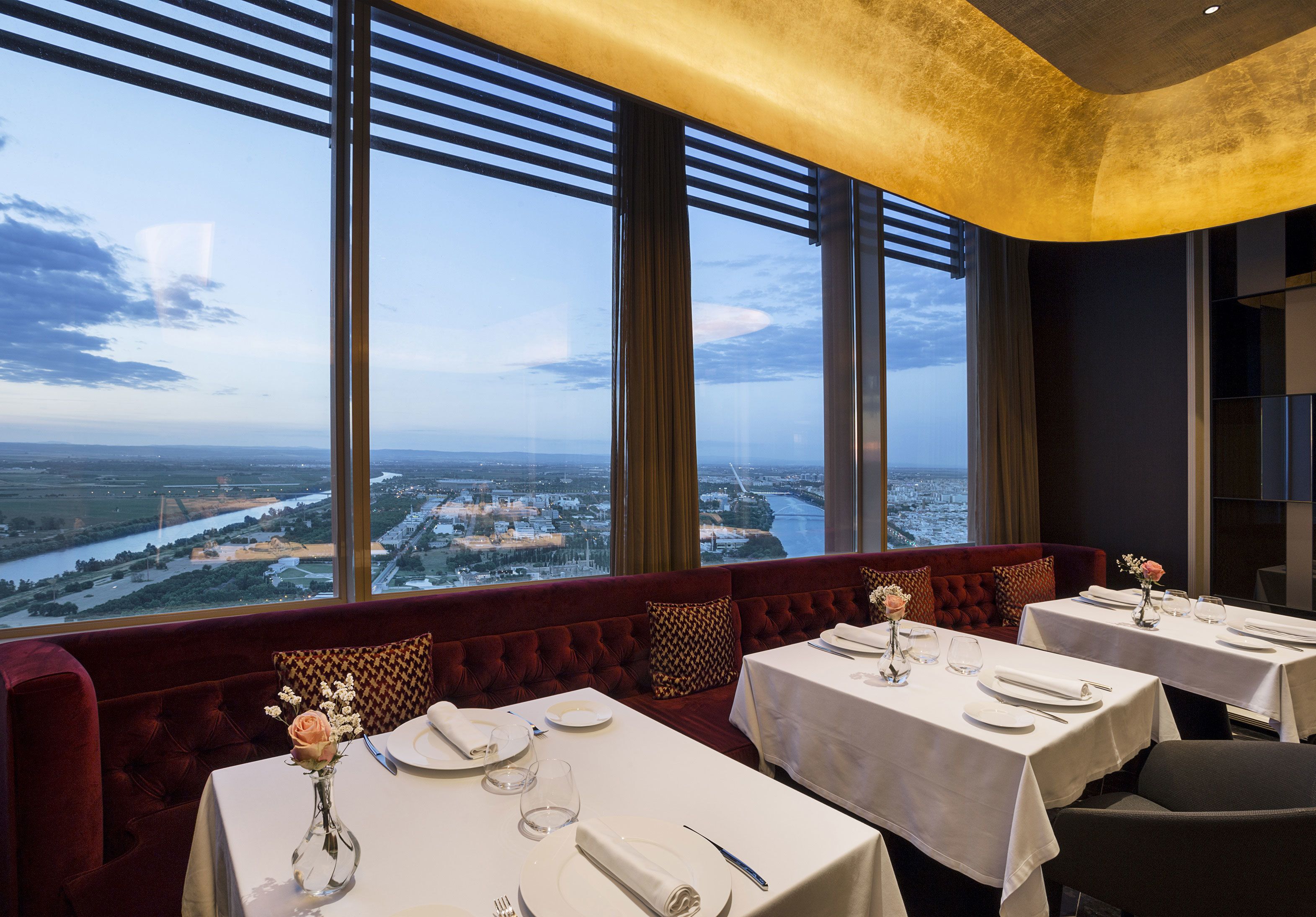 A restaurant with tables set for dining, large windows offering a view of a river and cityscape, and a red upholstered bench.