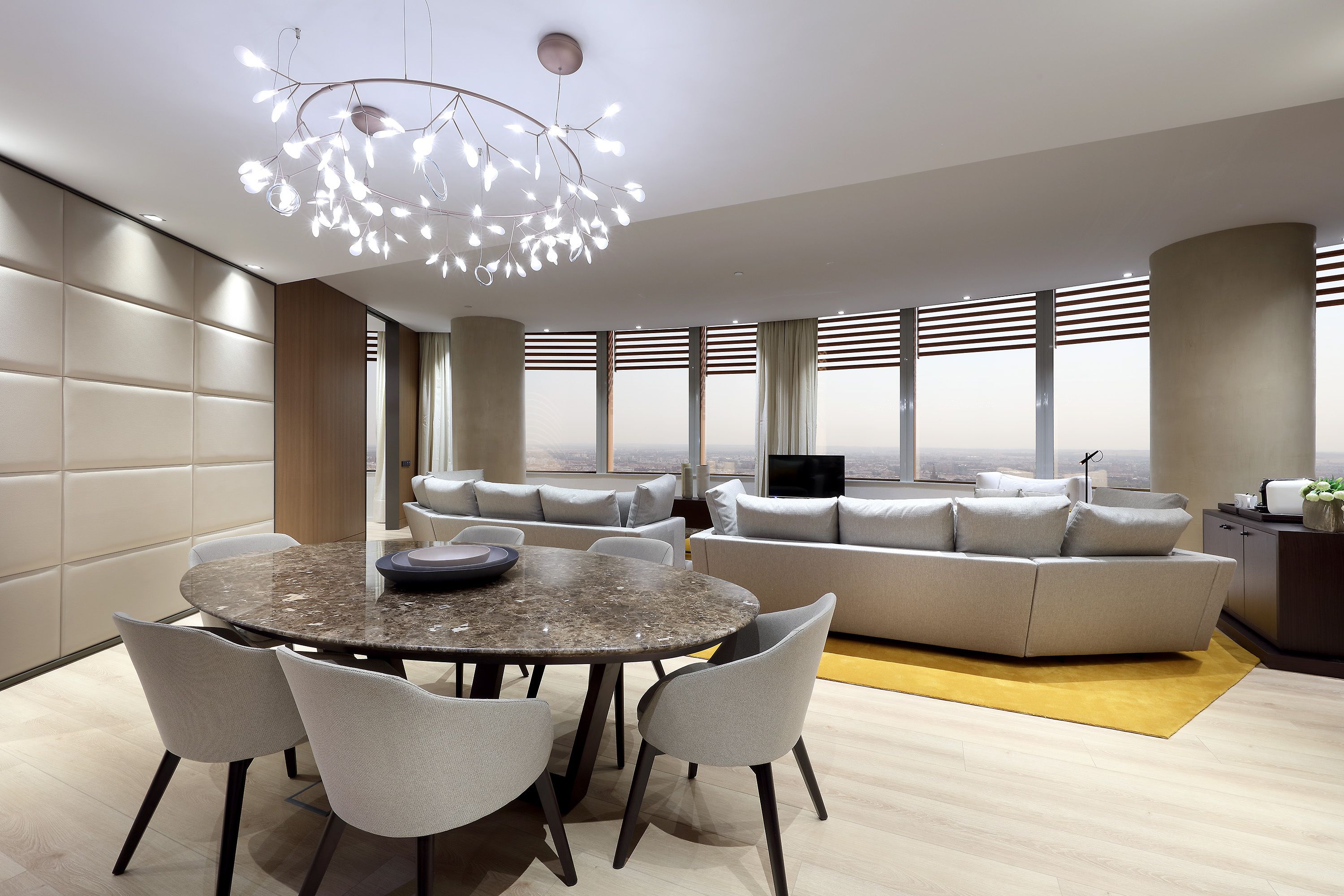 A modern living room with a round dining table, grey upholstered sofas, and a large window offering a city view.