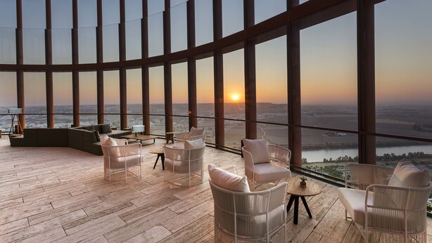A spacious balcony with a curved glass wall, furnished with white wicker chairs and a sofa, overlooking a sunset over a river. - Eurostars Torre Sevilla (Seville)