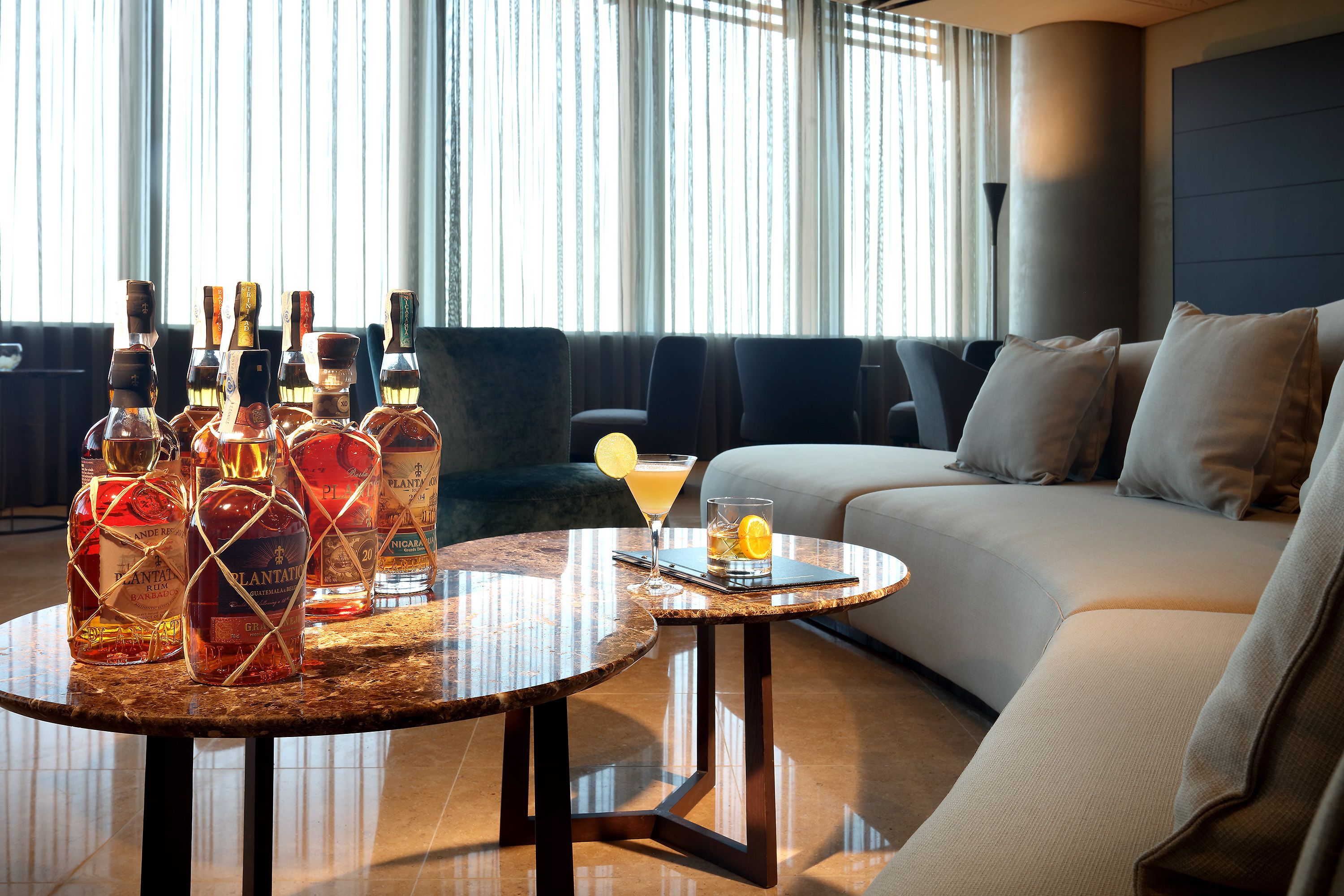 A modern living room with a round table displaying liquor bottles, a glass of cocktail, and a lemon slice. The room features a sofa and a large window with curtains.