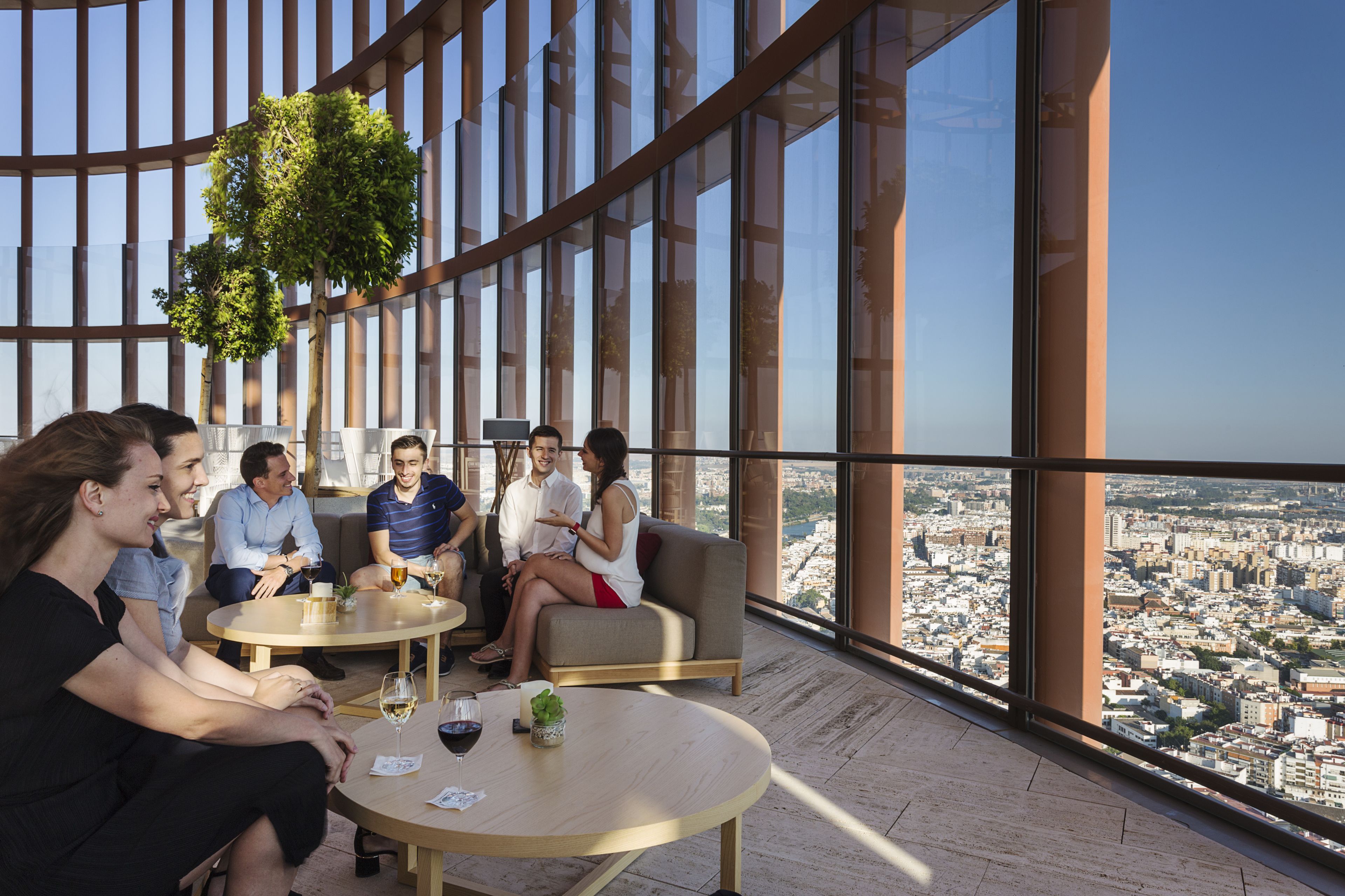 A group of people seated on a balcony with a city view, large windows, and a hanging plant.
