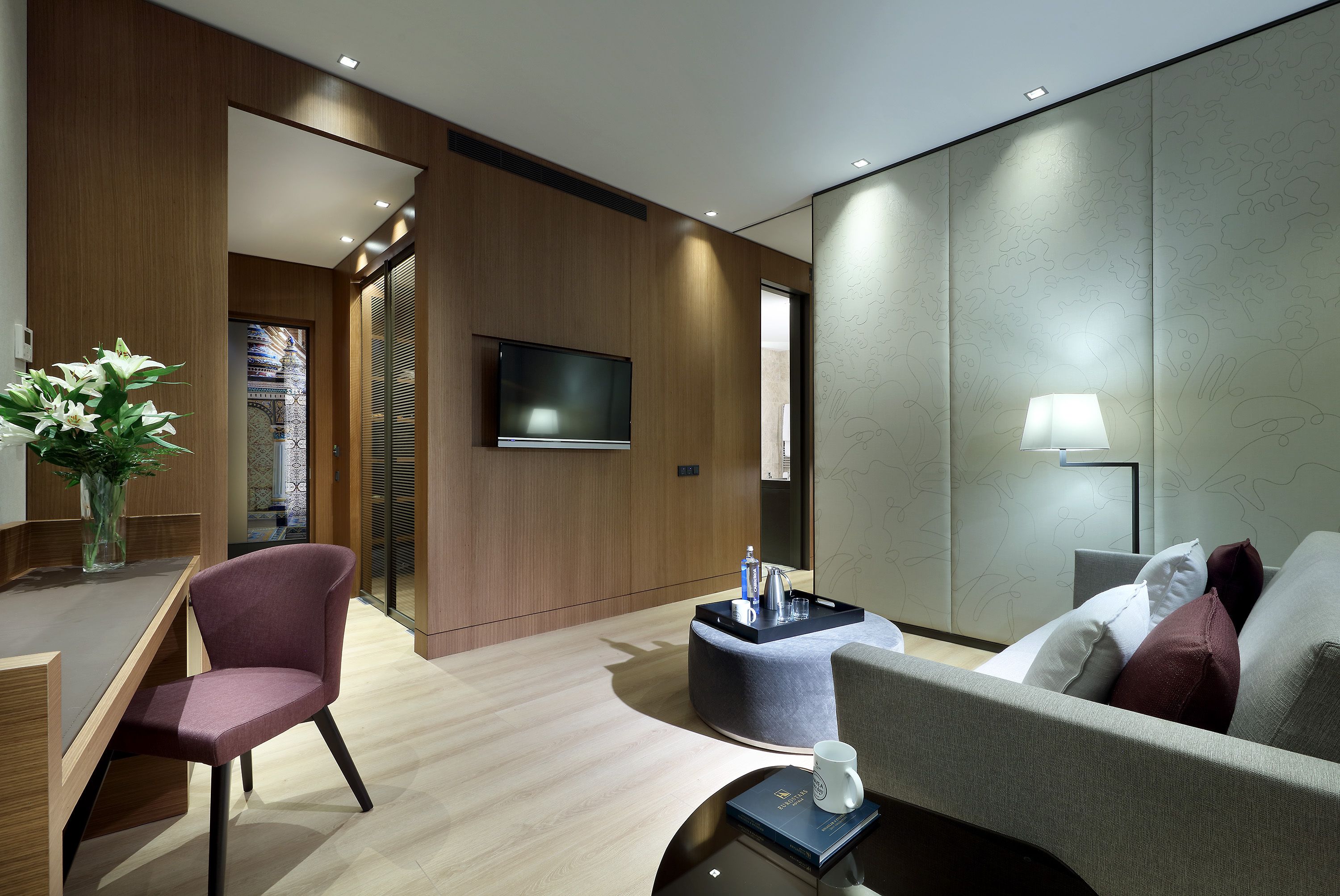 Una habitación de hotel moderna con suelo de madera, un televisor de pantalla plana montado en la pared, un otomán redondo con una lámpara, un sofá gris y un escritorio con una silla.