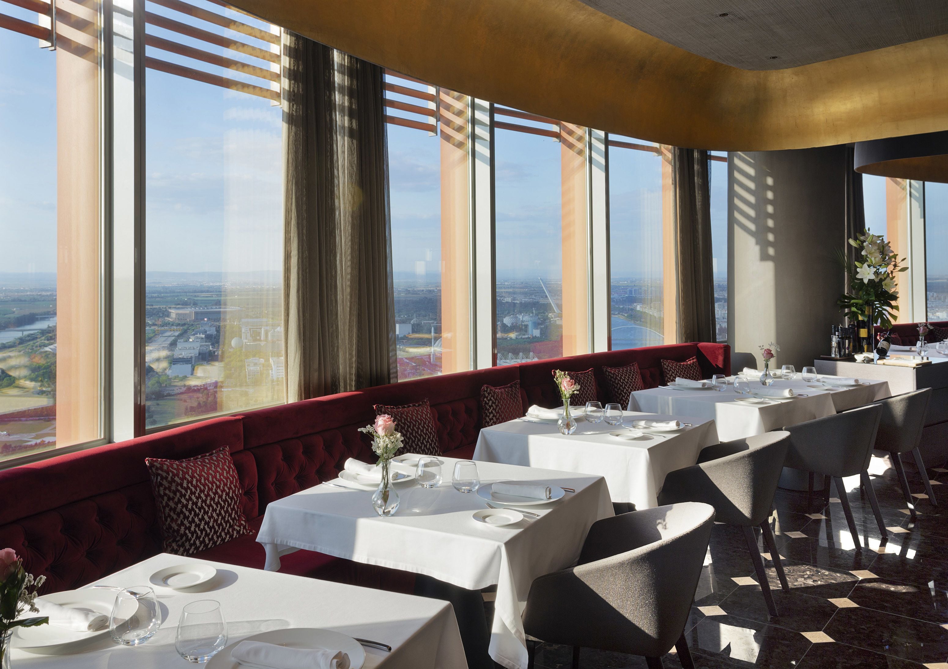 A restaurant with multiple tables set for dining, large windows offering a city view, and a red upholstered bench along the wall.