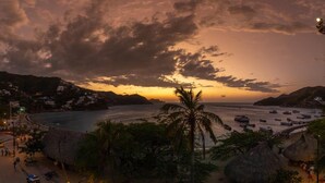 Un pueblo costero al atardecer con barcos en el agua, una playa con chozas de techo de paja y una montaña al fondo.