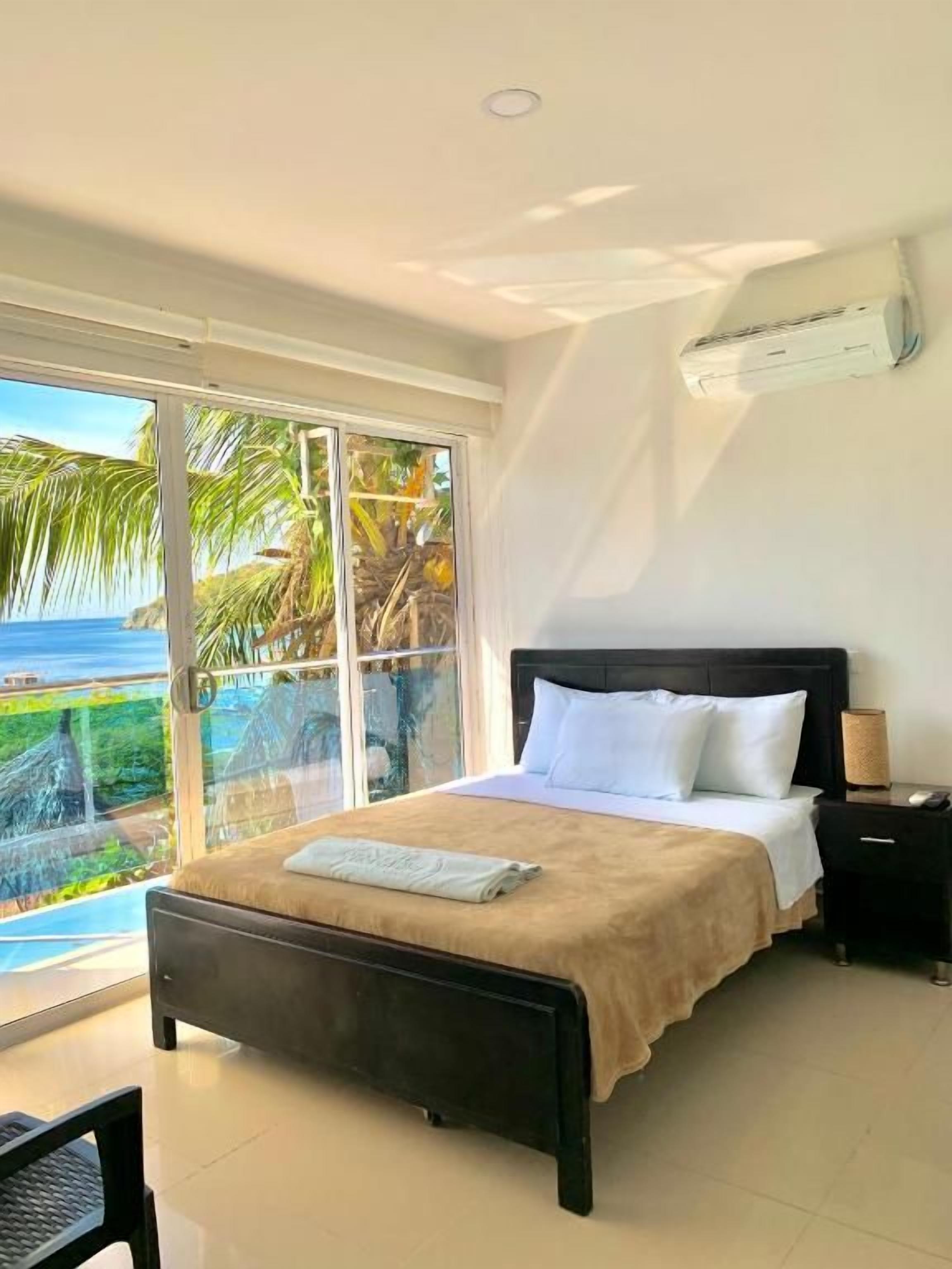 A bedroom with a bed, a nightstand, a lamp, and a view of the ocean.