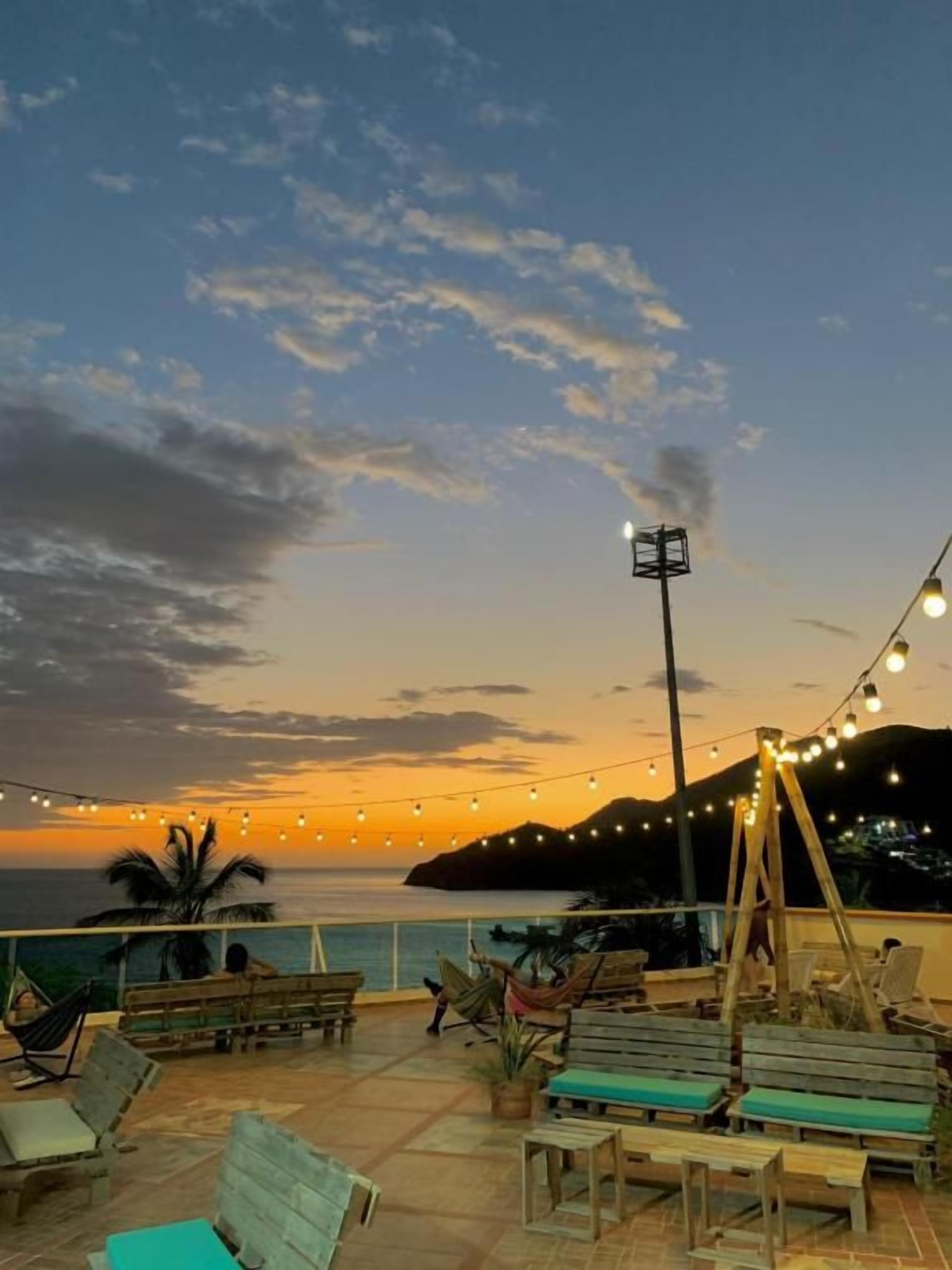 Un área de estar al aire libre con muebles de madera y luces colgantes frente a un atardecer.