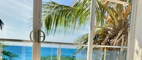 A room with a bed, a window with a view of the ocean, and a balcony with palm trees.
