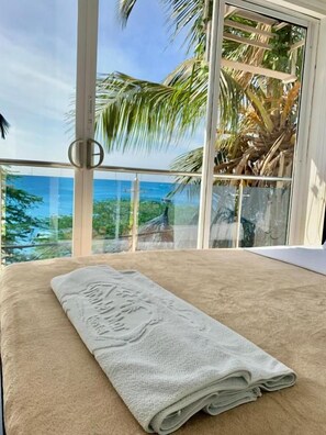 Una habitación con cama, ventana con vista al océano y balcón con palmeras.