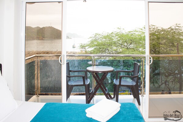 A balcony with a table and chairs, a bed with a blue towel, and a view of a body of water and trees. - Viña del Mar Hostal (Santa Marta)