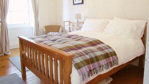 A bedroom with a wooden bed, a white headboard, a plaid blanket, a wicker chair, a small table with a lamp, and a framed picture on the wall. - Edinburgh City Retreat Apartments (Edinburgh)