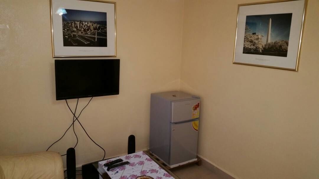Una habitación pequeña con televisión montada en la pared, un refrigerador y dos cuadros enmarcados.