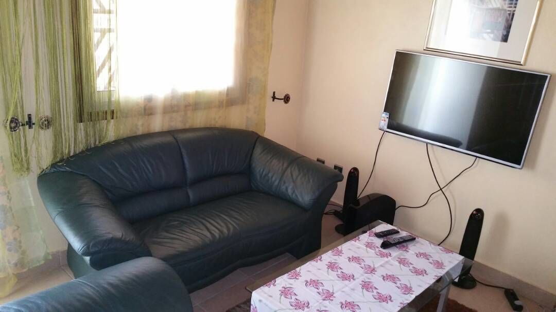Ett vardagsrum med en svart lädersoffa, ett litet bord med ett blommigt bordsduk och en platt-TV som är monterad på väggen.