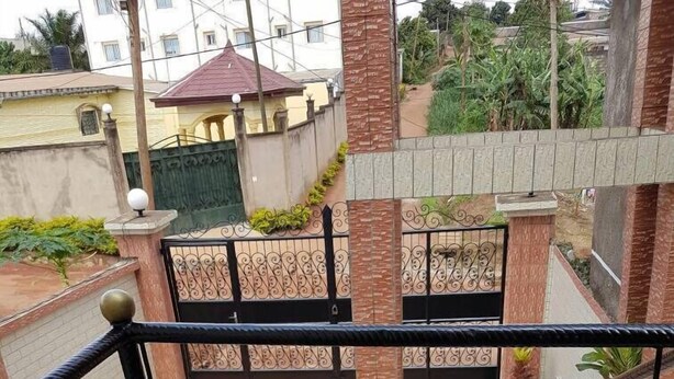 A balcony with a metal railing, potted plants, and a view of neighboring buildings and greenery. - Bs VIP Appartement (Yaoundé)