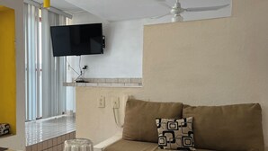 A living room with a sofa, a flat-screen TV mounted on the wall, a ceiling fan, and a window with blinds.