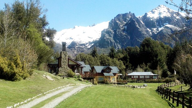 A mountain lodge with stone buildings, a gravel path, and snow-capped mountains in the background. - Tierra Sureña (San Carlos de Bariloche)
