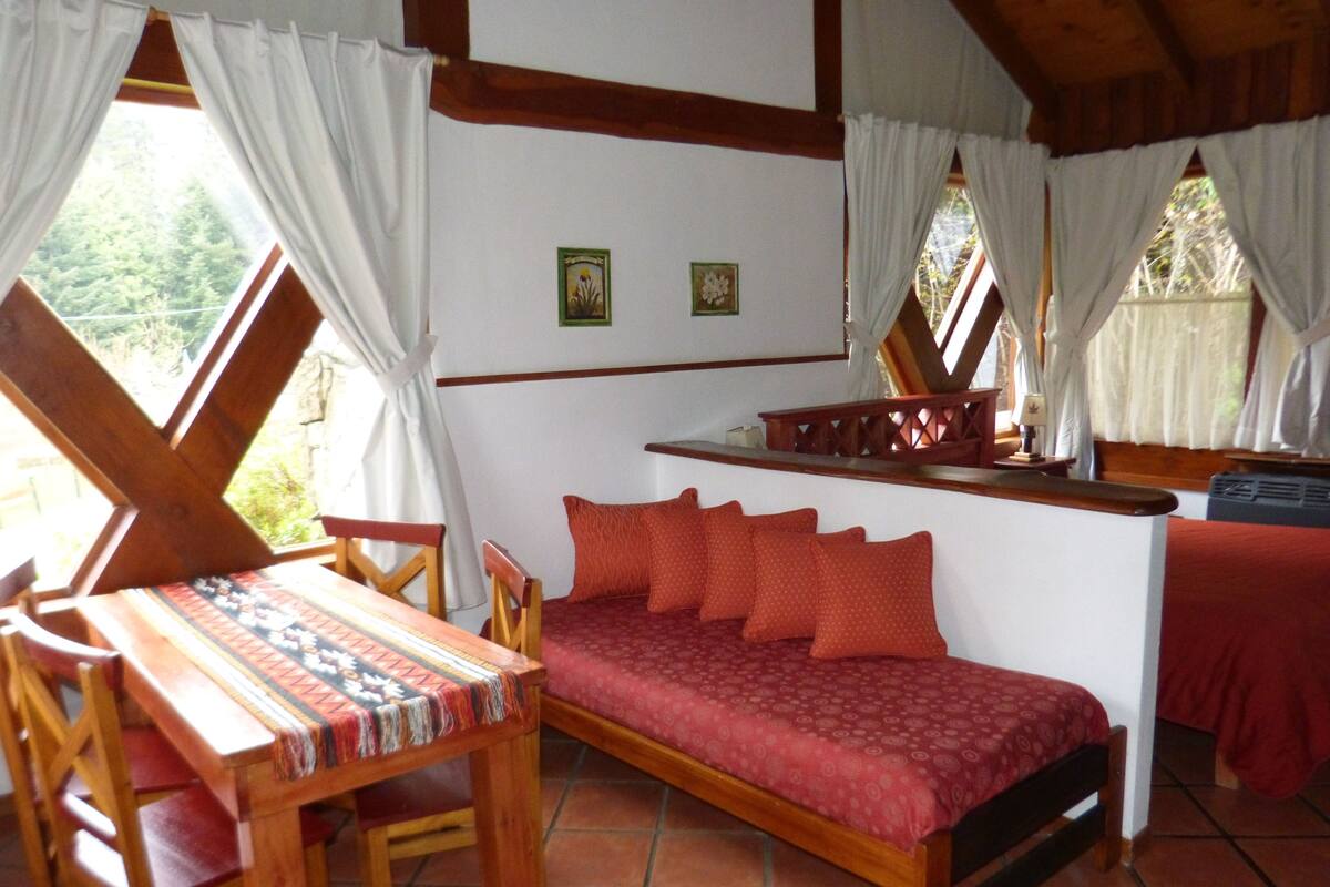 Une pièce chaleureuse avec un banc en bois, une table et un lit recouvert d’une literie rouge. Deux tableaux encadrés ornent le mur, et une fenêtre est agrémentée de rideaux.